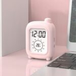 Réveil de voyage rose sur bureau rose avec plante verte, affichant l'heure et la température.