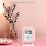 Réveil de voyage rose avec écran numérique affichant 12:00 et 28.6°C, posé près d'un ordinateur.
