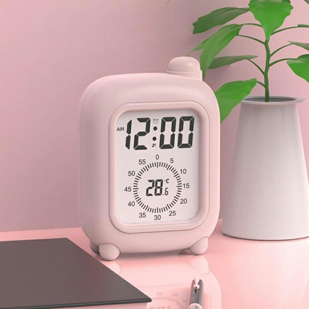 Réveil de voyage rose sur un bureau rose avec plante verte, affichage numérique et thermomètre intégré.