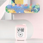 Réveil de voyage rose sur un bureau décoré, avec texte 'HELLO' et affichage de la température en anglais.