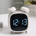 Réveil double cloche blanc avec affichage LED bleu, posé sur une table blanche.
